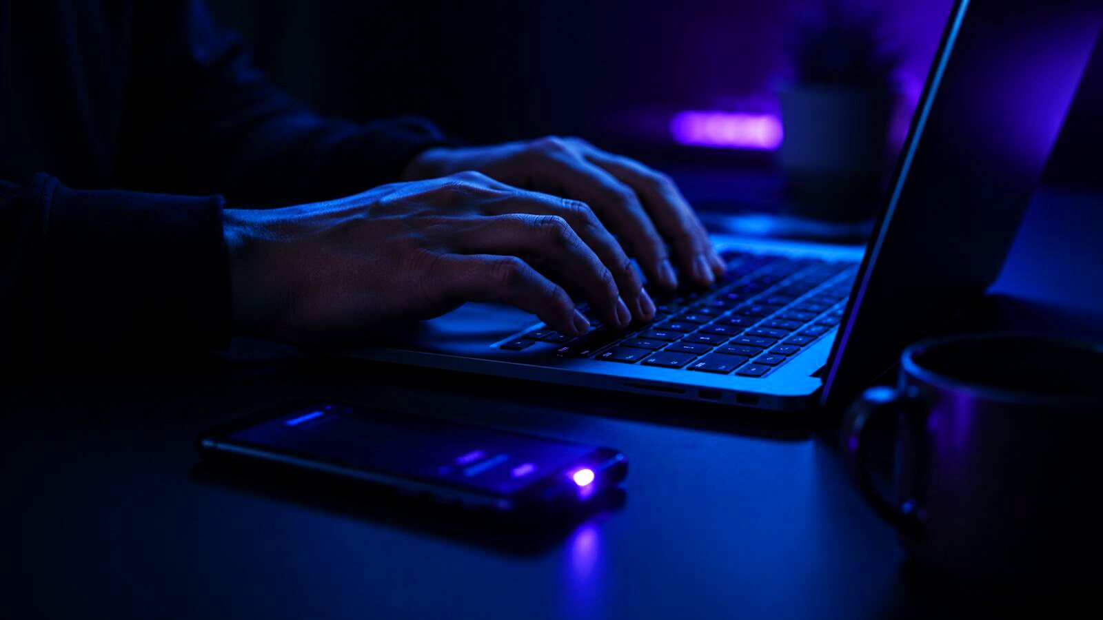 Close-up of hands typing on a laptop keyboard with a smartphone notification glowing nearby